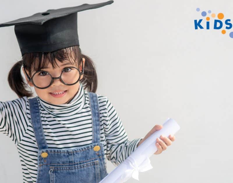 A little girl wearing a graduation cap beams with joy at KidsParks in Des Moines, WA, celebrating her accomplishments in a fun and vibrant setting.